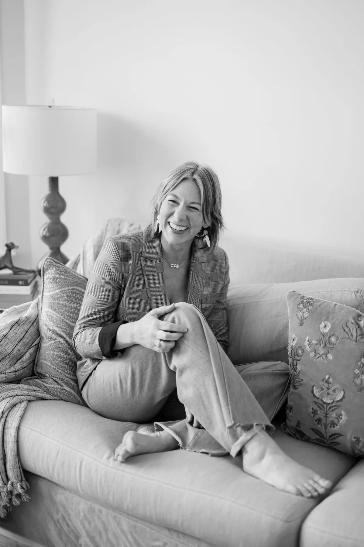 Tisha Leslie laughing, seated barefoot on a sofa in a plaid blazer.