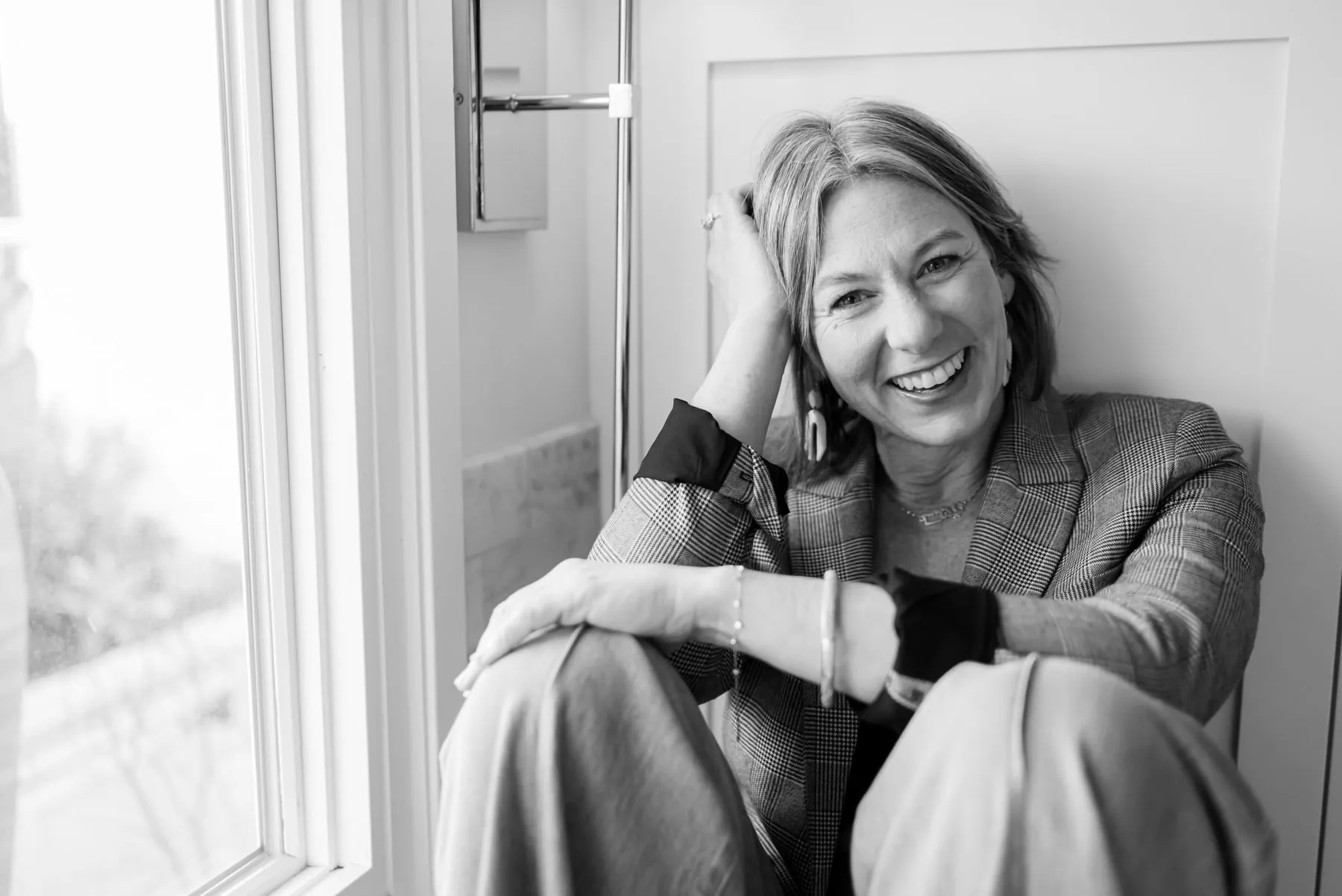 Tisha Leslie laughing beside a window, one hand in her hair, wearing a plaid blazer.